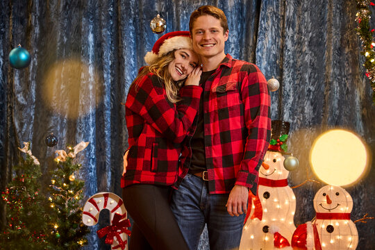 Couple wearing matching red buffalo plaid shirts pose together in festive Christmas photo studio setting