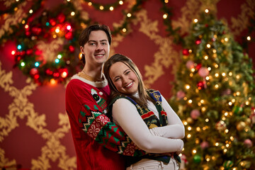 Happy couple wearing festive ugly Christmas sweaters posing in front of decorated holiday tree with twinkling lights