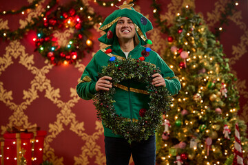 Man wearing festive Christmas tree costume holds decorated wreath in front of illuminated holiday trees