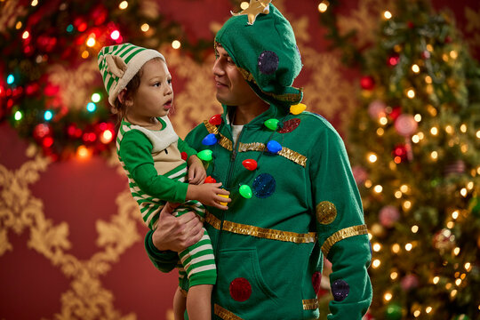 Father and child wear matching Christmas tree and elf costumes with festive holiday lights