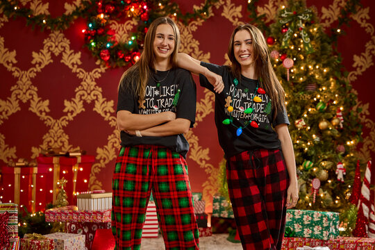 Two sisters wearing matching Christmas pajamas and graphic tees pose in front of decorated trees