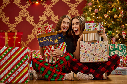 Two sisters in plaid pajamas holding wrapped Christmas gifts in front of decorated tree