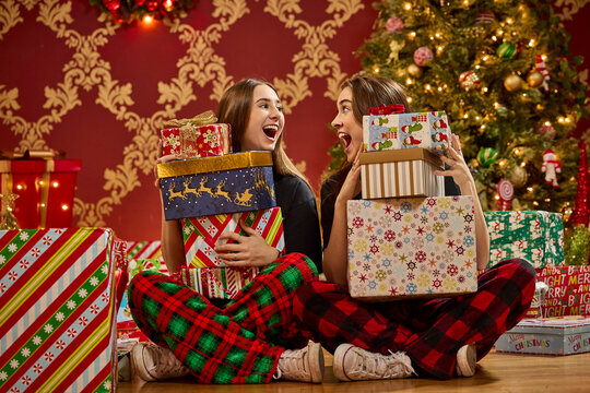 Two sisters in plaid pajamas holding stacks of wrapped Christmas presents by decorated tree