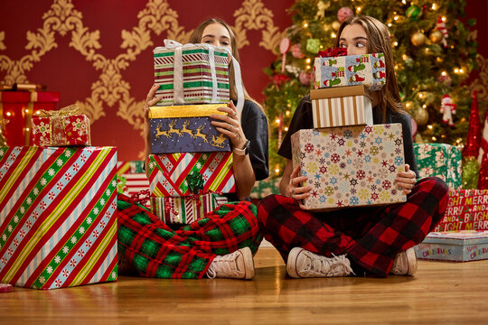 Two sisters in plaid pajamas holding stacks of wrapped Christmas presents near decorated tree