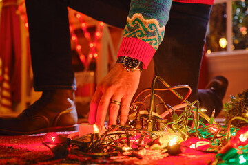 Person wearing festive sweater untangles colorful Christmas lights on table with holiday decorations