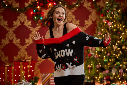 Woman showing off festive ugly Christmas sweater with snow theme and holiday decorations in background
