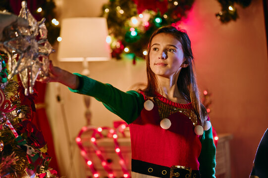 Young girl wearing festive elf sweater decorates Christmas tree with silver star topper at home