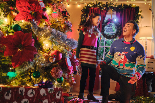 Couple decorating Christmas tree together in festive sweaters with holiday lights and ornaments at home