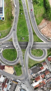 Imagem a&eacute;rea em movimento mostrando rotat&oacute;ria urbana com m&uacute;ltiplas faixas e ve&iacute;culos em circula&ccedil;&atilde;o constante, destacando a din&acirc;mica do tr&aacute;fego e a organiza&ccedil;&atilde;o da infraestrutura vi&aacute;ria