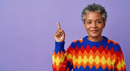 Confident middle-aged African American woman with grey hair smiling and pointing up, wearing a vibrant colorful patterned sweater on a purple background.
