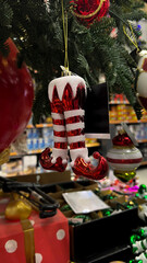 Vertical mobile photo. Glass New Year toy, elf red pants, Santa shoes on Christmas tree branch in supermarket, mass market store. Concept of Merry Christmas eve, shopping, gift, sale, winter holiday