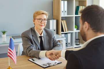 Handshake seals business agreement between client and consultant. Man sitting in office of US public services or embassy having consular visa interview with officer with US flag at desk