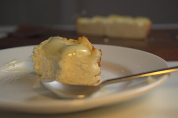 appetizing slice of homemade cottage cheese casserole topped with flowing condensed milk on white plate with fork bite and whole casserole in soft background