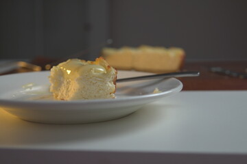 appetizing slice of homemade cottage cheese casserole topped with flowing condensed milk on white plate with fork bite and whole casserole in soft background