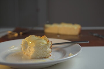 appetizing slice of homemade cottage cheese casserole topped with flowing condensed milk on white plate with fork bite and whole casserole in soft background