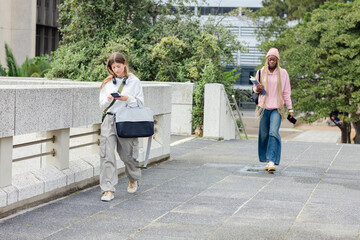 Diverse female students walking at campus courtyard carrying notebooks and checking smartphones