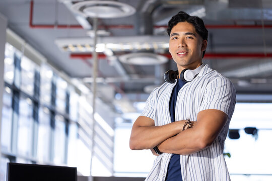 Asian man standing at right in open-plan office wearing headphones near monitor, copy space
