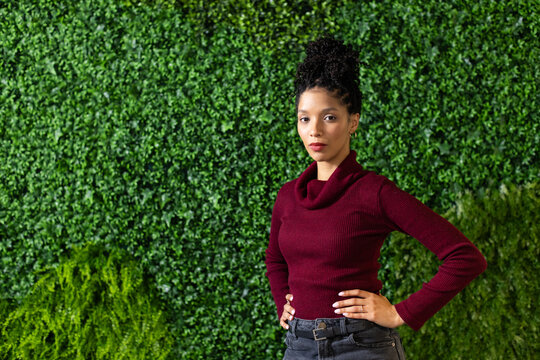 African American woman wearing burgundy knit sweater and jeans posing before plant wall, copy space