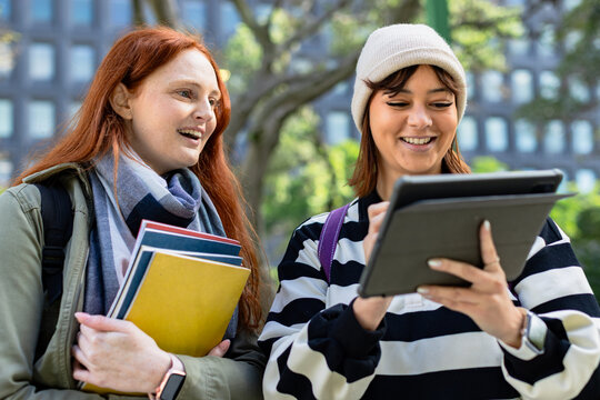 Two female students collaborating on campus walkway, holding notebooks and using stylus tablet