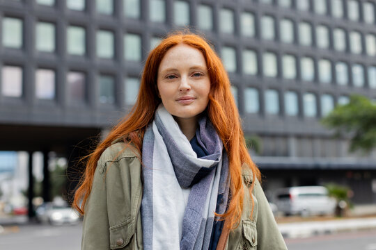 Woman standing on city sidewalk wearing olive green utility jacket and striped scarf beneath clouds