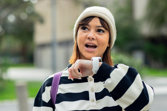 Woman wearing white beanie, striped top checking smartwatch carrying backpack strap on campus path