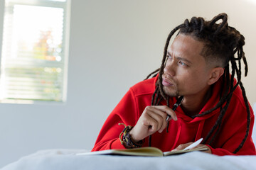 Mid adult African American man in red hoodie leaning in bedroom under blinds, writing in notebook