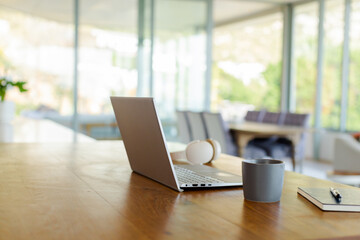 Laptop computer resting on wooden desk with headphones, coffee mug, notebook and pen near windows