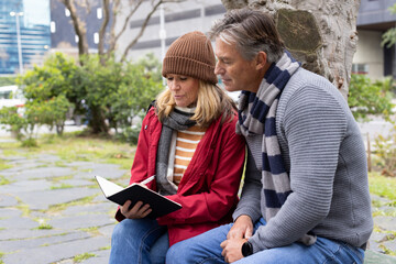 Senior couple reading open notebook on stone bench by tree trunk in park wearing red jacket