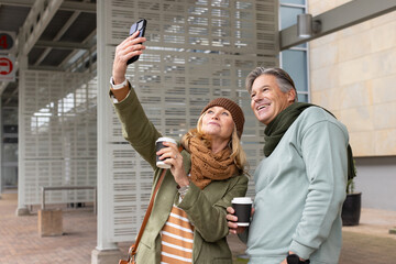 Senior couple taking selfie under canopy on transit platform holding smartphone and two coffee cups