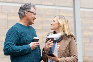 Senior couple standing on sidewalk in front of stone facade holding coffee cups and phone chatting