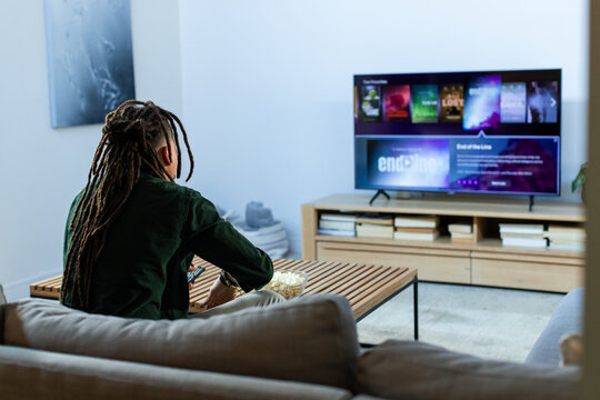 Person with dreads sitting on sofa facing mounted TV holding popcorn bowl, remote in living room