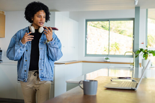 African American man holding smartphone speaking on headset near laptop, mug in kitchen, copy space - Powered by Adobe