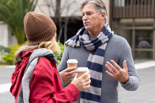 Middle-aged friends wearing scarves and beanie standing in courtyard holding coffee cups, gesturing