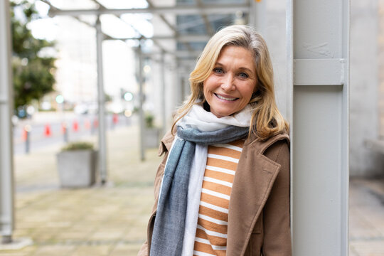 Senior woman leaning against column on sidewalk under scaffolding near cones wearing coat and scarf