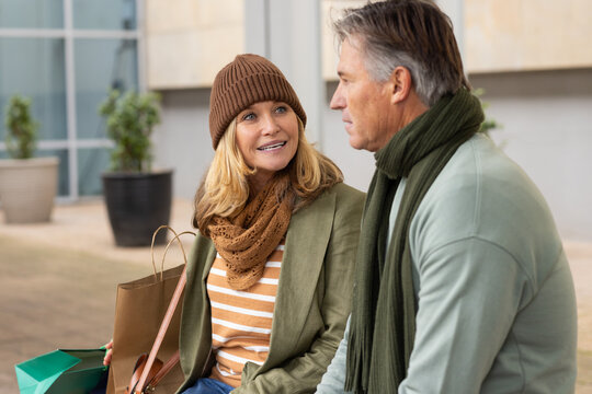 Senior couple sitting on wooden bench outside modern shopping plaza, holding leather and paper bags