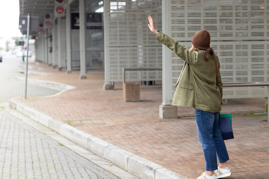 Woman raising arm and hailing bus at station platform with canvas tote bag and knit beanie