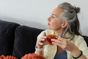Senior woman sitting on dark sofa holding teacup of tea while gazing right, copy space