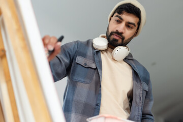 Indian man wearing beanie and headphones applying pink paint at wooden easel with tray in studio