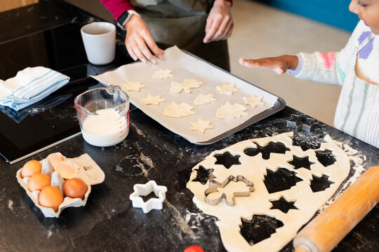 Mother and daughter baking cookies on countertop in home kitchen with rolling pin, cookie cutters