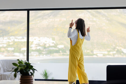 Woman standing at home in yellow overalls with headphones pointing toward windows overlooking sea