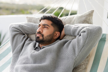 Adult South Asian man reclining in striped hammock on terrace with grey pillow, ropes and railing