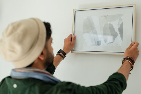 Asian man wearing smartwatch mounting framed abstract art on white wall at home, copy space