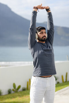 Indian man stretching arms overhead on seaside terrace wearing over-ear headphones and bracelets