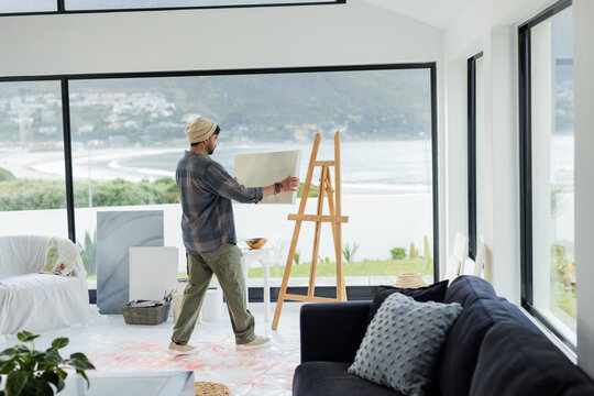 Male artist is holding blank canvas before wooden easel in coastal studio overlooking calm sea