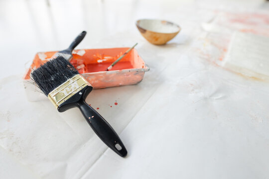 Paintbrush is lying across white drop cloth, with paint tray, stir stick, bowl, orange splatters