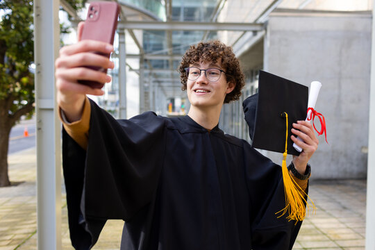 Male wearing graduation gown on campus path holding cap, diploma raising smartphone taking selfie