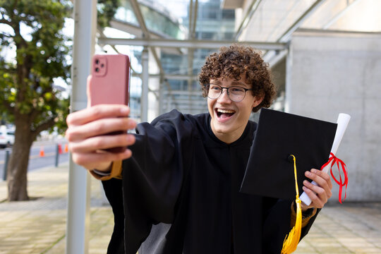 Male graduate taking selfie using pink smartphone with mortarboard cap and diploma scroll on campus
