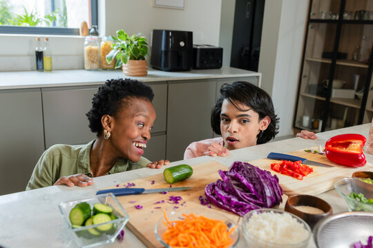 Diverse friends leaning over kitchen counter, peering at chopped vegetables on wooden cutting board