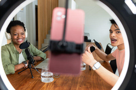 Diverse coworkers speaking into microphones at office table with ring light and smartphone