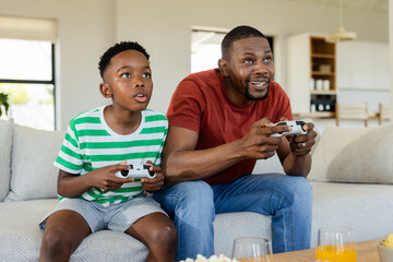 African American father and son sitting on sofa in living room playing game with white controllers